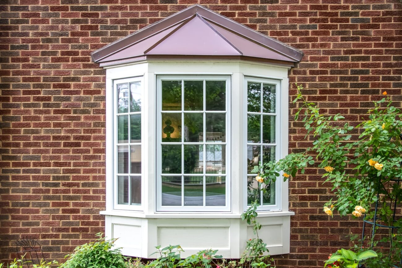 Bay window with copper roof on traditional brick home with flowers below and yellow roses extending out onto the window in springtime