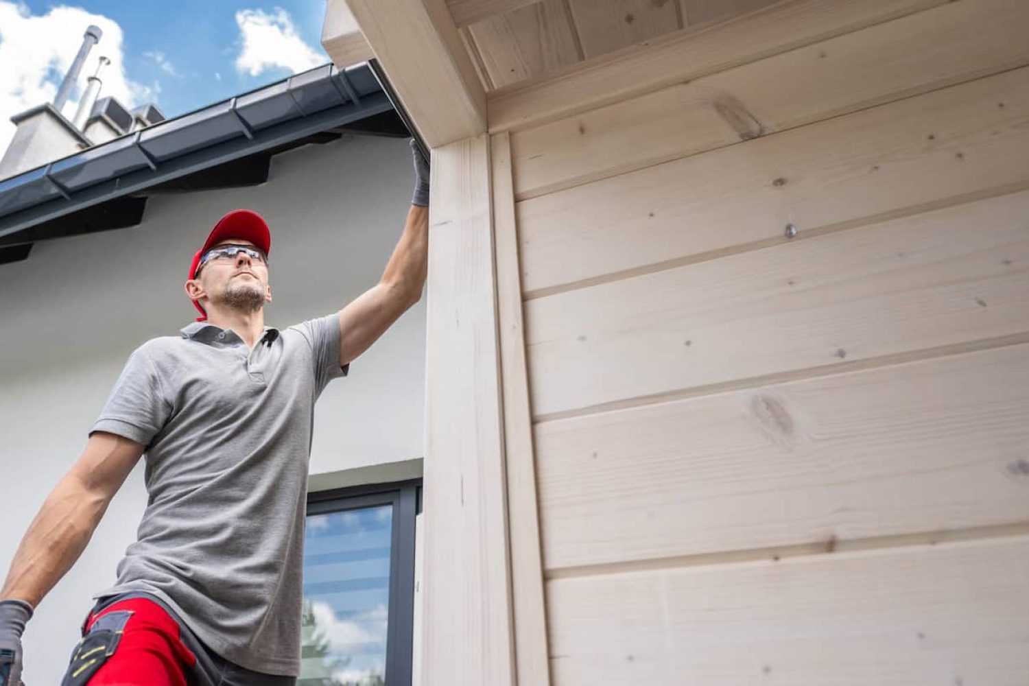 A person is inspecting and working on the wooden exterior of a house under a clear blue sky.