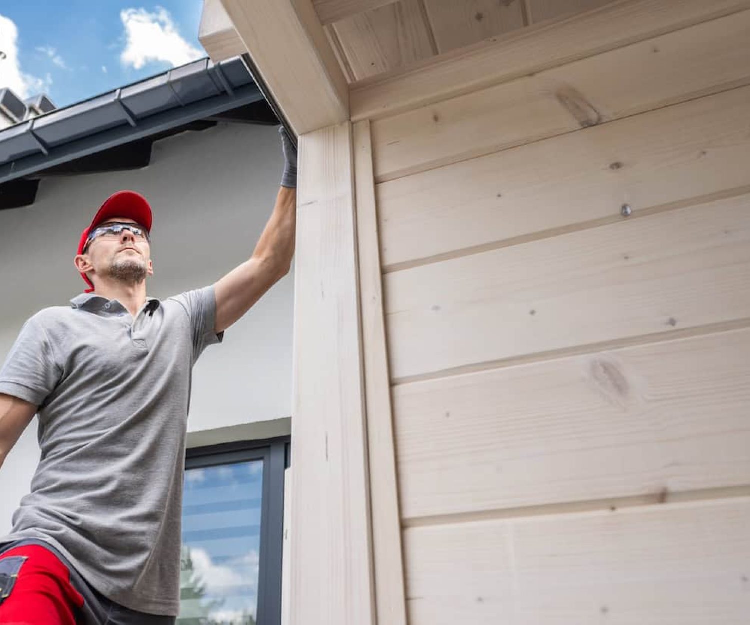 A person is inspecting and working on the wooden exterior of a house under a clear blue sky.